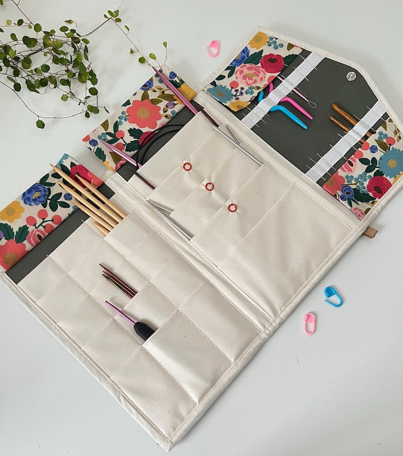 Pode incluir: Um estojo de tecido para organizar agulhas de tric&ocirc; e croch&ecirc; com um padr&atilde;o floral de flores rosas, azuis e amarelas sobre um fundo creme. O interior cont&eacute;m v&aacute;rios bolsos com diversas agulhas e ganchos de metal e madeira.