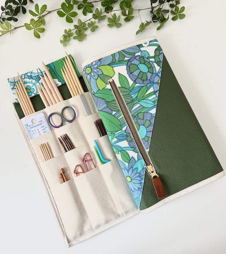 Pode incluir: Um estojo de tecido para agulhas de tric&ocirc; com um padr&atilde;o floral em tons de azul e verde, combinado com uma sec&ccedil;&atilde;o em verde-azeitona s&oacute;lido. O interior cont&eacute;m agulhas de madeira, agulhas de croch&eacute; de metal, marcadores e um calibrador.