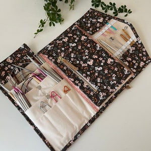 Pode incluir: Um estojo organizador de tecido preto com um pequeno padr&atilde;o floral rosa e branco, apresentando v&aacute;rios bolsos e compartimentos internos. Cont&eacute;m v&aacute;rias agulhas de tric&ocirc; de metal, agulhas circulares, agulhas de croch&ecirc; e alfinetes grandes.