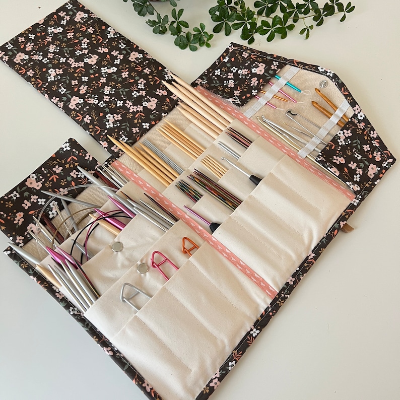Pode incluir: Um estojo de tecido para organizar agulhas de tric&ocirc; e croch&ecirc; com uma estampa floral castanho-escura de pequenas flores cor-de-rosa e brancas. O interior &eacute; forrado com bolsos de tecido creme contendo v&aacute;rias agulhas de metal e madeira.