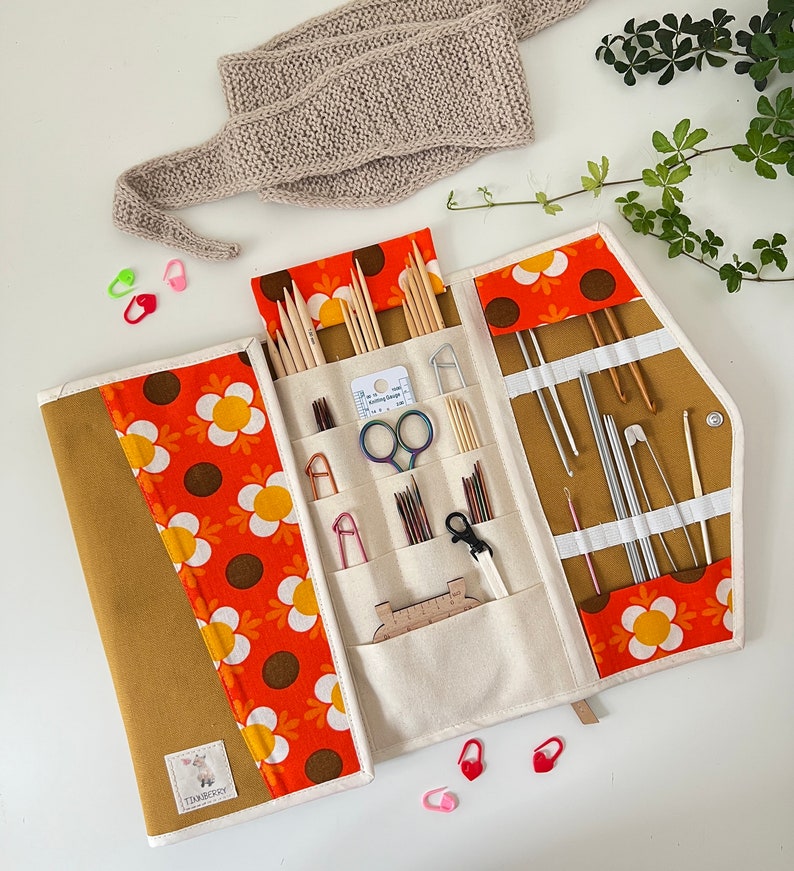 Pode incluir: Um estojo de tecido para agulhas de tric&ocirc; e croch&ecirc; com um padr&atilde;o floral retro em laranja, amarelo e castanho. O interior cont&eacute;m v&aacute;rios bolsos com agulhas de madeira e metal, agulhas de croch&ecirc;, tesouras e um pequeno medidor de agulhas.