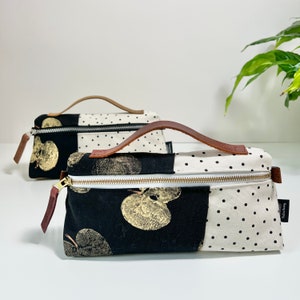 May include: Two black and white fabric zippered pouches with brown leather handles. One pouch has a black and white polka dot pattern and the other has a black background with a gold leaf print. Both pouches have a gold zipper.