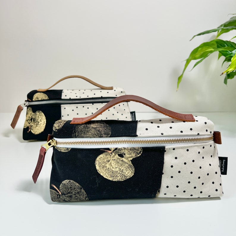May include: Two black and white fabric zippered pouches with brown leather handles. One pouch has a black and white polka dot pattern and the other has a black background with a gold leaf print. Both pouches have a gold zipper.