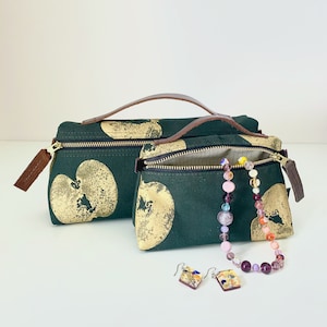 May include: Two green fabric zippered pouches with gold apple print and brown leather handles. A colorful beaded necklace and two small glass earrings are in front of the pouches.
