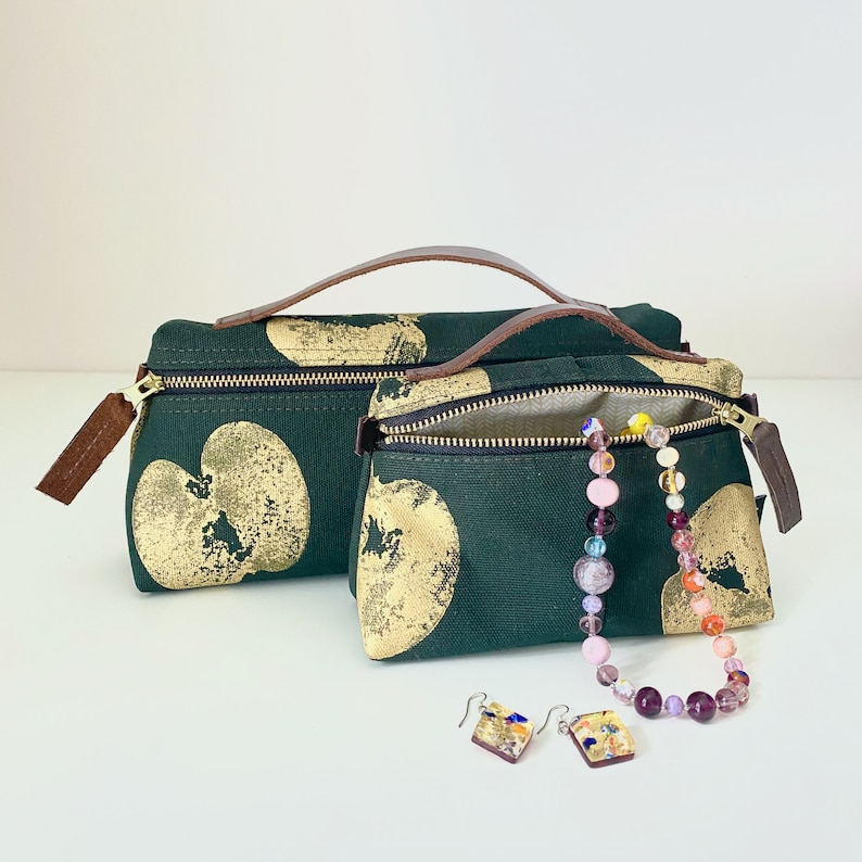 May include: Two green fabric zippered pouches with gold apple print and brown leather handles. A colorful beaded necklace and two small glass earrings are in front of the pouches.