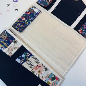 Pode incluir: Um estojo organizador de tecido com interior na cor creme, apresentando m&uacute;ltiplos bolsos e el&aacute;sticos. O exterior e os detalhes internos s&atilde;o feitos de tecido azul-marinho e tecido com estampa floral colorida. Uma etiqueta exibe TINKBERRY.
