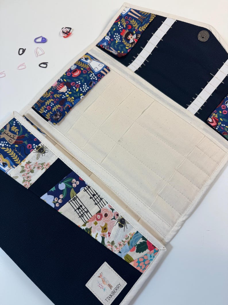 Pode incluir: Um estojo organizador de tecido com interior na cor creme, apresentando m&uacute;ltiplos bolsos e el&aacute;sticos. O exterior e os detalhes internos s&atilde;o feitos de tecido azul-marinho e tecido com estampa floral colorida. Uma etiqueta exibe TINKBERRY.
