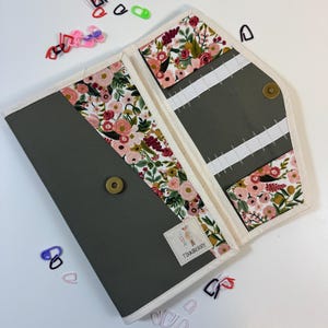 Pode incluir: Um estojo organizador de tecido cinza com padr&atilde;o floral de flores rosas, vermelhas e verdes. O estojo possui acabamento branco, fecho de press&atilde;o em lat&atilde;o e el&aacute;sticos internos. Uma pequena etiqueta branca com o texto TIANBERRY est&aacute; fixada.