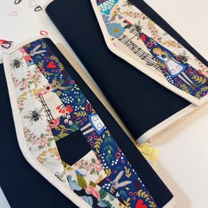 Pode incluir: Dois estojos de tecido azul-marinho com uma aba decorativa em design patchwork. O padr&atilde;o inclui ilustra&ccedil;&otilde;es de uma menina, abelhas, flores, cora&ccedil;&otilde;es e notas musicais sobre um fundo creme e azul escuro com bordas brancas.