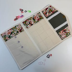 Pode incluir: Um estojo organizador de tecido com um padr&atilde;o floral de flores cor-de-rosa, vermelhas e verdes sobre um fundo branco. O interior tem v&aacute;rios bolsos de cor creme e uma sec&ccedil;&atilde;o cinzento-escura com presilhas el&aacute;sticas brancas, fechado com um bot&atilde;o de press&atilde;o.
