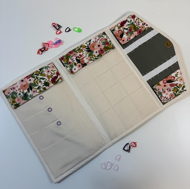 Pode incluir: Um estojo organizador de tecido com um padr&atilde;o floral de flores cor-de-rosa, vermelhas e verdes sobre um fundo branco. O interior tem v&aacute;rios bolsos de cor creme e uma sec&ccedil;&atilde;o cinzento-escura com presilhas el&aacute;sticas brancas, fechado com um bot&atilde;o de press&atilde;o.