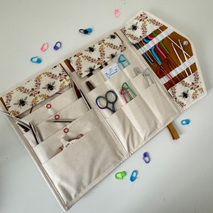 Pode incluir: Um estojo organizador de tecido com estampa de flores e abelhas, contendo v&aacute;rias ferramentas de tric&ocirc; e croch&ecirc;. O interior inclui bolsos com agulhas de metal, ganchos, uma pequena tesoura iridescente, um medidor de agulhas e marcadores de pontos de pl&aacute;stico coloridos espalhados ao redor do estojo aberto.