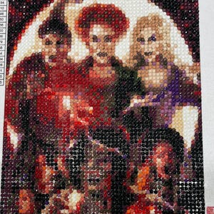 May include: Diamond painting of three figures with red, orange, and purple tones. The figures are set against a white background. The artwork is composed of many small, colorful, round pieces. The image is a close-up of the finished artwork.