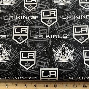 May include: Black and white fabric with a repeating pattern of the LA Kings logo. The logo features a crown, the letters LA, and the words LA Kings. The fabric is perfect for crafting projects, such as pillows, blankets, and tote bags.