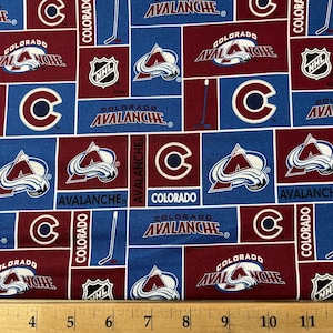 May include: A red and blue fabric with a repeating pattern of the Colorado Avalanche hockey team logo. The logo features a mountain range with a 'C' in the center and the words 'Colorado Avalanche' on either side. The fabric also includes the NHL logo and hockey sticks.