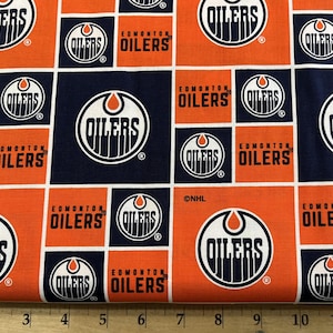 May include: Orange and navy blue fabric with a repeating pattern of the Edmonton Oilers hockey team logo. The logo features a white circle with a white drop inside, the words "Oilers" in white, and a white ring around the circle. The fabric also includes the words "Edmonton Oilers" in white.