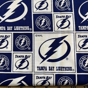May include: White fabric with a repeating pattern of the Tampa Bay Lightning hockey team logo. The logo is a blue lightning bolt inside a white circle with the words "Tampa Bay Lightning" and "Hockey Club" in blue text. The background is a dark blue color.