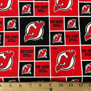 May include: Red and black fabric with a repeating pattern of the New Jersey Devils NHL hockey team logo. The logo is a white and red stylized "NJ" inside a circle with a red devil's tail.