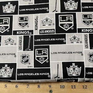May include: Black and white fabric with a repeating pattern of the Los Angeles Kings hockey team logo, including the team's name, the NHL logo, and a crown. The fabric is 14 inches wide.