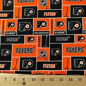 May include: Orange and black fabric with a repeating pattern of the Philadelphia Flyers logo, the team name, and a hockey stick. The fabric is 17 inches wide.