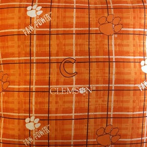 May include: An orange fabric featuring a plaid pattern with thin black and white lines. The design includes repeating white paw prints, the text PAW POWER, a large letter C, and the word CLEMSON with a paw print replacing the letter O.