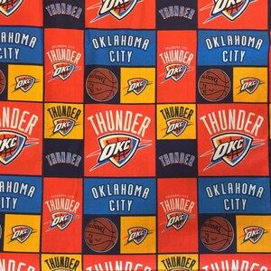 May include: A fabric with a repeating pattern of Oklahoma City Thunder logos and text. The design features the team's logo, basketballs, and the words "Oklahoma City" and "Thunder" in red, blue, and yellow squares.