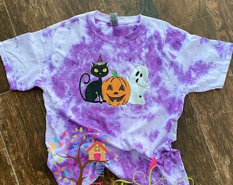 May include: A purple tie-dye t-shirt featuring a Halloween graphic. The graphic includes a black cat wearing a witch hat, a jack-o'-lantern, and a white ghost. The text "The Treehouse" is visible at the bottom of the shirt.