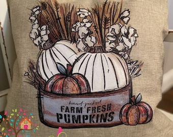 May include: A beige square pillow with a fall-themed design. The pillow features a graphic of white pumpkins, wheat, and cotton, with the text "hand-picked FARM FRESH PUMPKINS" on a metal container. A small pumpkin is on the right.