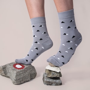 May include: A pair of grey socks with a black and white triangle pattern. The socks are being worn by a person standing on a stack of grey rocks. The top rock has a red circle painted on it.