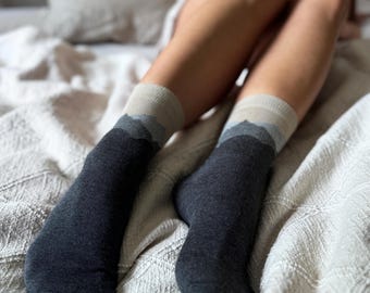 Puede incluir: Un par de calcetines grises oscuros con un diseño de montaña en tonos grises y beige en el puño. Los calcetines se llevan en las piernas de una persona, descansando sobre una manta texturizada de color blanco roto.