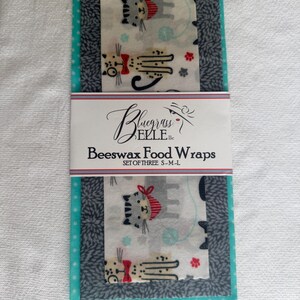 May include: A set of three beeswax food wraps featuring a white fabric with a cat pattern, a dark gray fabric with a leaf-like pattern, and a teal fabric with white polka dots. A paper band reads Bluegrass ELLE llc Beeswax Food Wraps SET OF THREE S-M-L.
