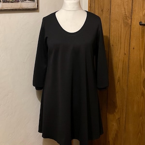 May include: A black dress with a scoop neckline and three-quarter length sleeves. The dress has a loose, flowing shape and is displayed on a mannequin. The fabric is solid and opaque. The dress is a simple, elegant design.