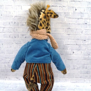 Puede incluir: Un peluche de jirafa hecho a mano, visto desde atr&aacute;s, con una chaqueta azul, pantalones a rayas y una bufanda beige. La jirafa tiene una cabeza manchada y una melena de piel sint&eacute;tica. El juguete est&aacute; sobre una pared de ladrillos blancos.