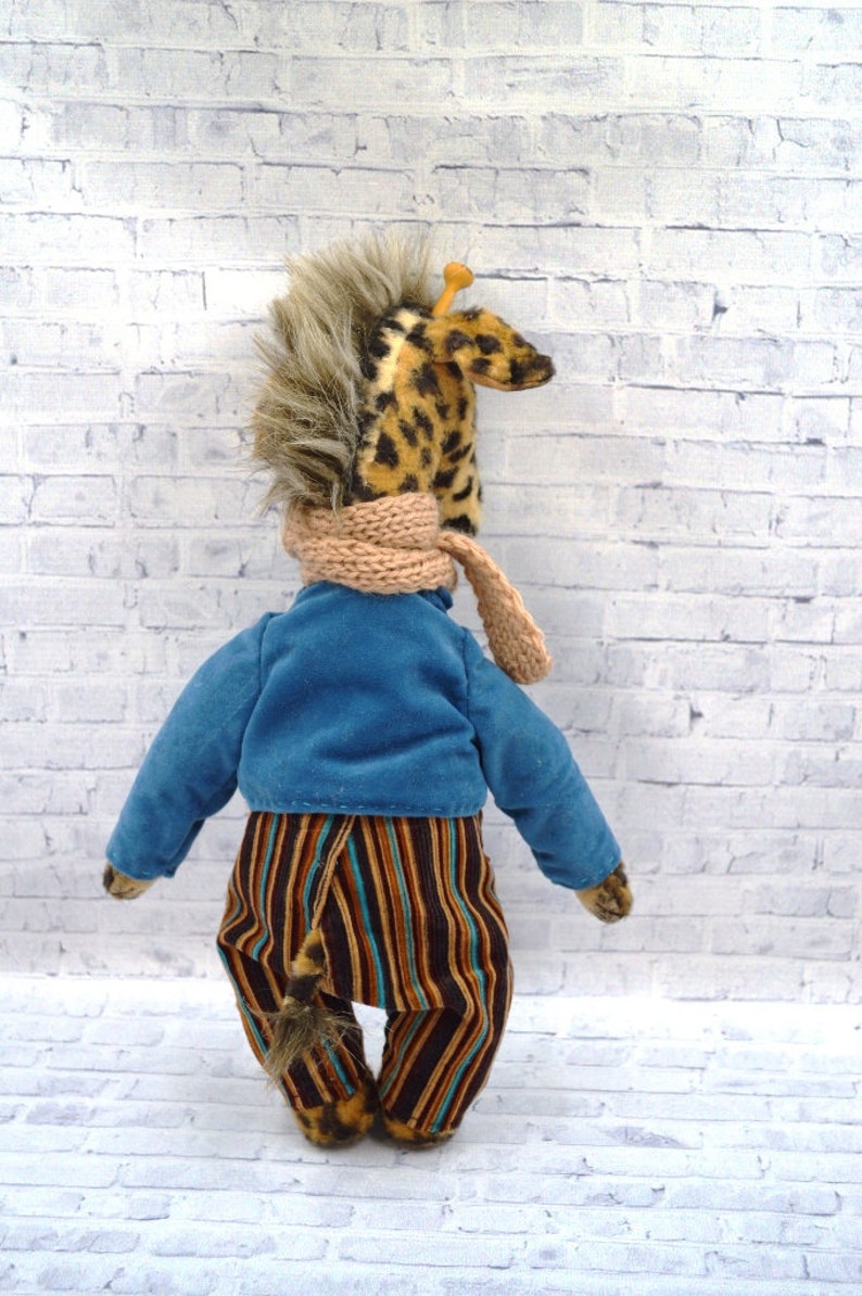 Puede incluir: Un peluche de jirafa hecho a mano, visto desde atr&aacute;s, con una chaqueta azul, pantalones a rayas y una bufanda beige. La jirafa tiene una cabeza manchada y una melena de piel sint&eacute;tica. El juguete est&aacute; sobre una pared de ladrillos blancos.