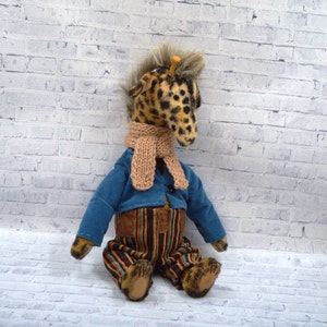Puede incluir: Un peluche de jirafa hecho a mano con cabeza manchada, que lleva una chaqueta azul, pantalones a rayas y una bufanda beige. La jirafa tiene una melena esponjosa y est&aacute; sentada contra una pared de ladrillos blancos.