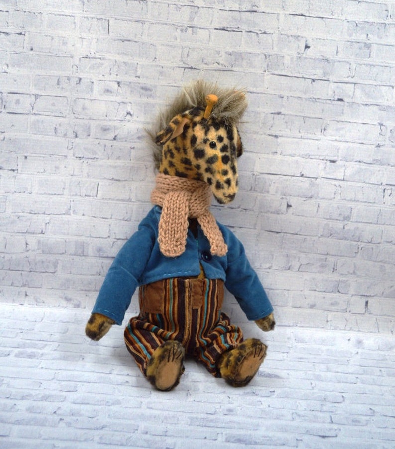 Puede incluir: Un peluche de jirafa hecho a mano con cabeza manchada, que lleva una chaqueta azul, pantalones a rayas y una bufanda beige. La jirafa tiene una melena esponjosa y est&aacute; sentada contra una pared de ladrillos blancos.