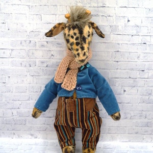 Puede incluir: Un peluche de jirafa con la cabeza manchada, que lleva una chaqueta azul, pantalones a rayas y una bufanda beige. La jirafa tiene peque&ntilde;os cuernos y est&aacute; colocada contra una pared de ladrillos blancos.