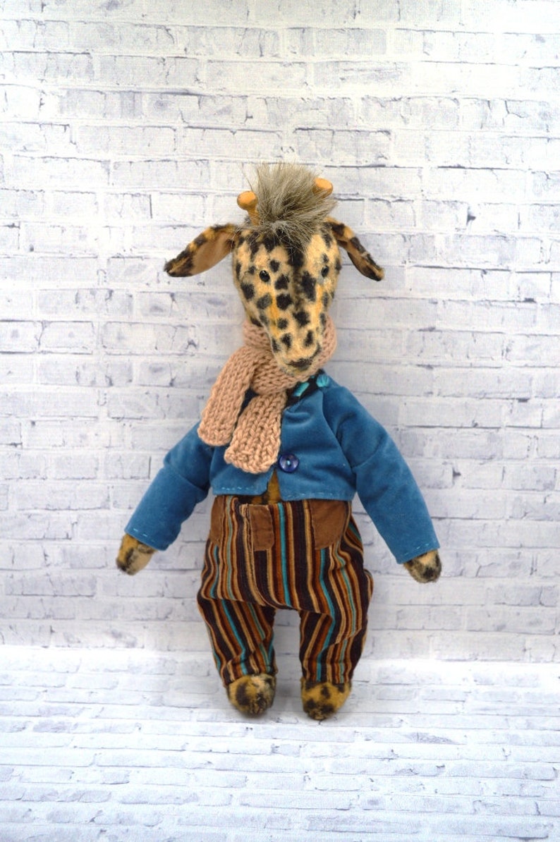 Puede incluir: Un peluche de jirafa con la cabeza manchada, que lleva una chaqueta azul, pantalones a rayas y una bufanda beige. La jirafa tiene peque&ntilde;os cuernos y est&aacute; colocada contra una pared de ladrillos blancos.