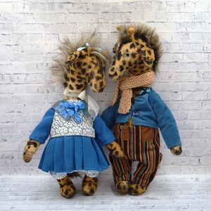 Puede incluir: Dos peluches de jirafa hechos a mano. Uno lleva un vestido azul con un top de encaje blanco y un lazo azul. La otra jirafa lleva una chaqueta azul, pantalones a rayas y una bufanda color canela. Ambos tienen pelaje manchado.