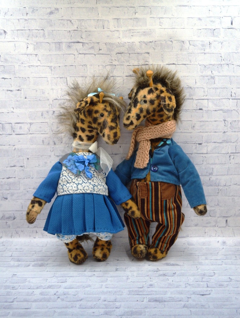 Puede incluir: Dos peluches de jirafa hechos a mano. Uno lleva un vestido azul con un top de encaje blanco y un lazo azul. La otra jirafa lleva una chaqueta azul, pantalones a rayas y una bufanda color canela. Ambos tienen pelaje manchado.
