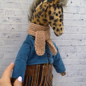 Puede incluir: Un peluche de jirafa con cabeza manchada, que lleva una chaqueta azul, pantalones marrones y a rayas, y una bufanda beige. La jirafa tiene una melena de piel sint&eacute;tica y est&aacute; de pie frente a una pared de ladrillos blancos.