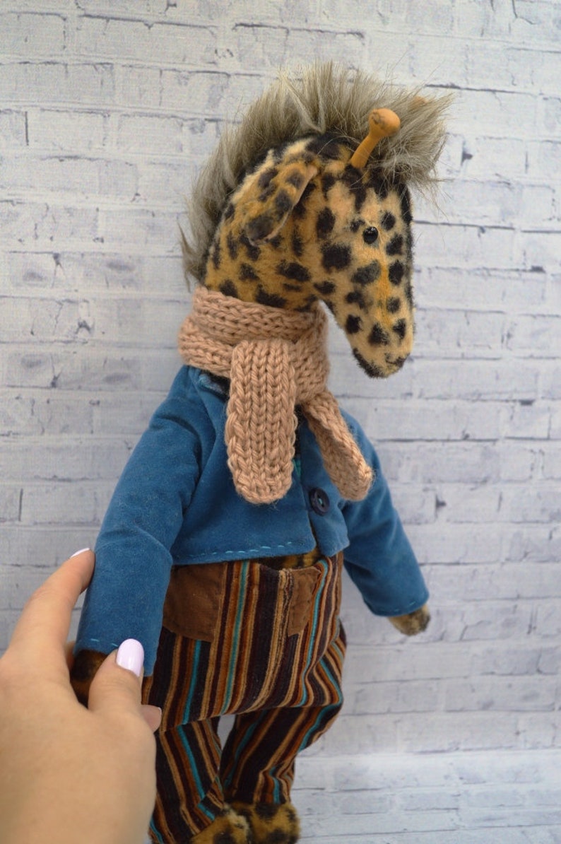 Puede incluir: Un peluche de jirafa con cabeza manchada, que lleva una chaqueta azul, pantalones marrones y a rayas, y una bufanda beige. La jirafa tiene una melena de piel sint&eacute;tica y est&aacute; de pie frente a una pared de ladrillos blancos.