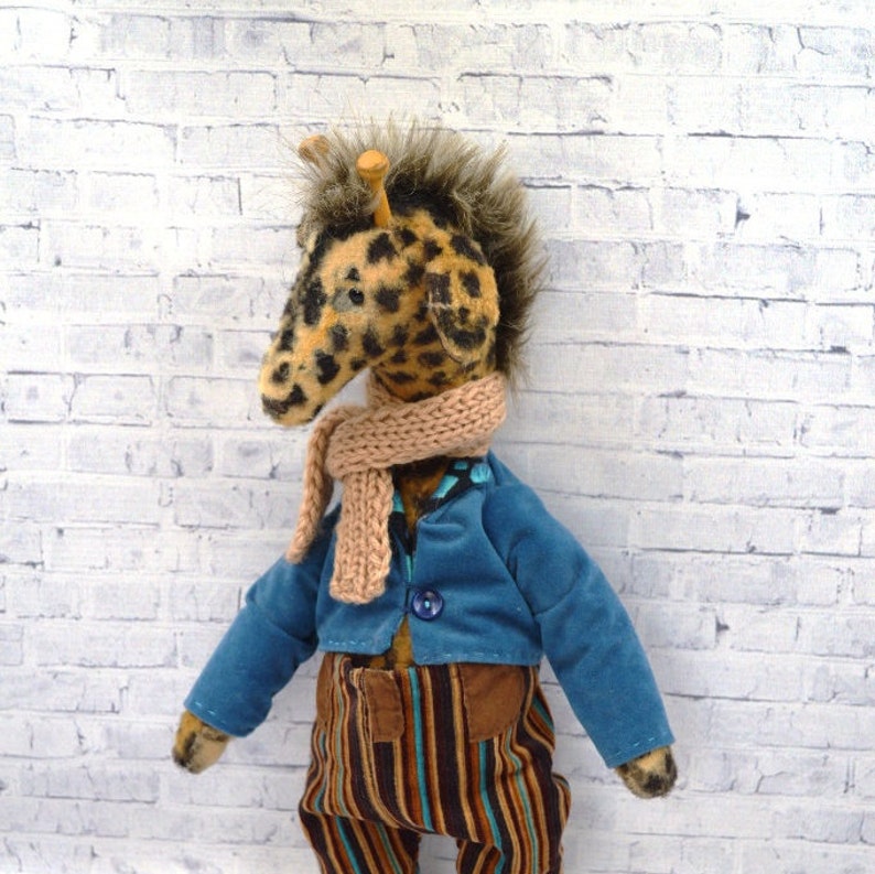 Puede incluir: Un juguete de peluche de jirafa con cabeza manchada, que lleva una chaqueta azul, pantalones a rayas y una bufanda beige. La jirafa tiene una melena esponjosa y peque&ntilde;os cuernos de madera. Los pantalones tienen bolsillos marrones.