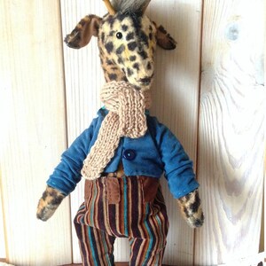 Puede incluir: Un peluche de jirafa hecho a mano con un patr&oacute;n de manchas, que lleva una chaqueta azul, pantalones a rayas y una bufanda beige. La jirafa tiene peque&ntilde;os cuernos de madera y un mech&oacute;n de pelo gris.