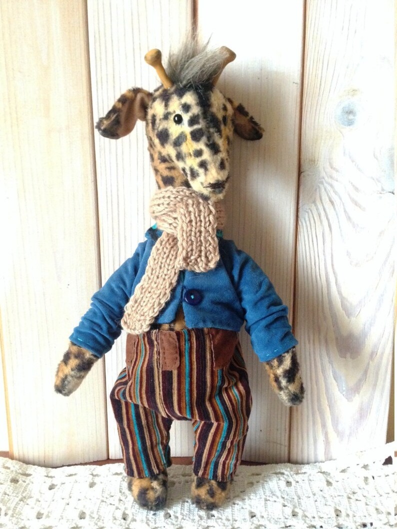 Puede incluir: Un peluche de jirafa hecho a mano con un patr&oacute;n de manchas, que lleva una chaqueta azul, pantalones a rayas y una bufanda beige. La jirafa tiene peque&ntilde;os cuernos de madera y un mech&oacute;n de pelo gris.