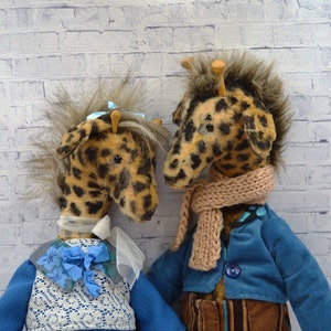 Puede incluir: Dos mu&ntilde;ecos de peluche de jirafa con pelaje moteado, uno frente al otro. Uno lleva un vestido azul con un chaleco de encaje blanco y una flor azul. El otro lleva una chaqueta azul, pantalones a rayas y una bufanda beige.