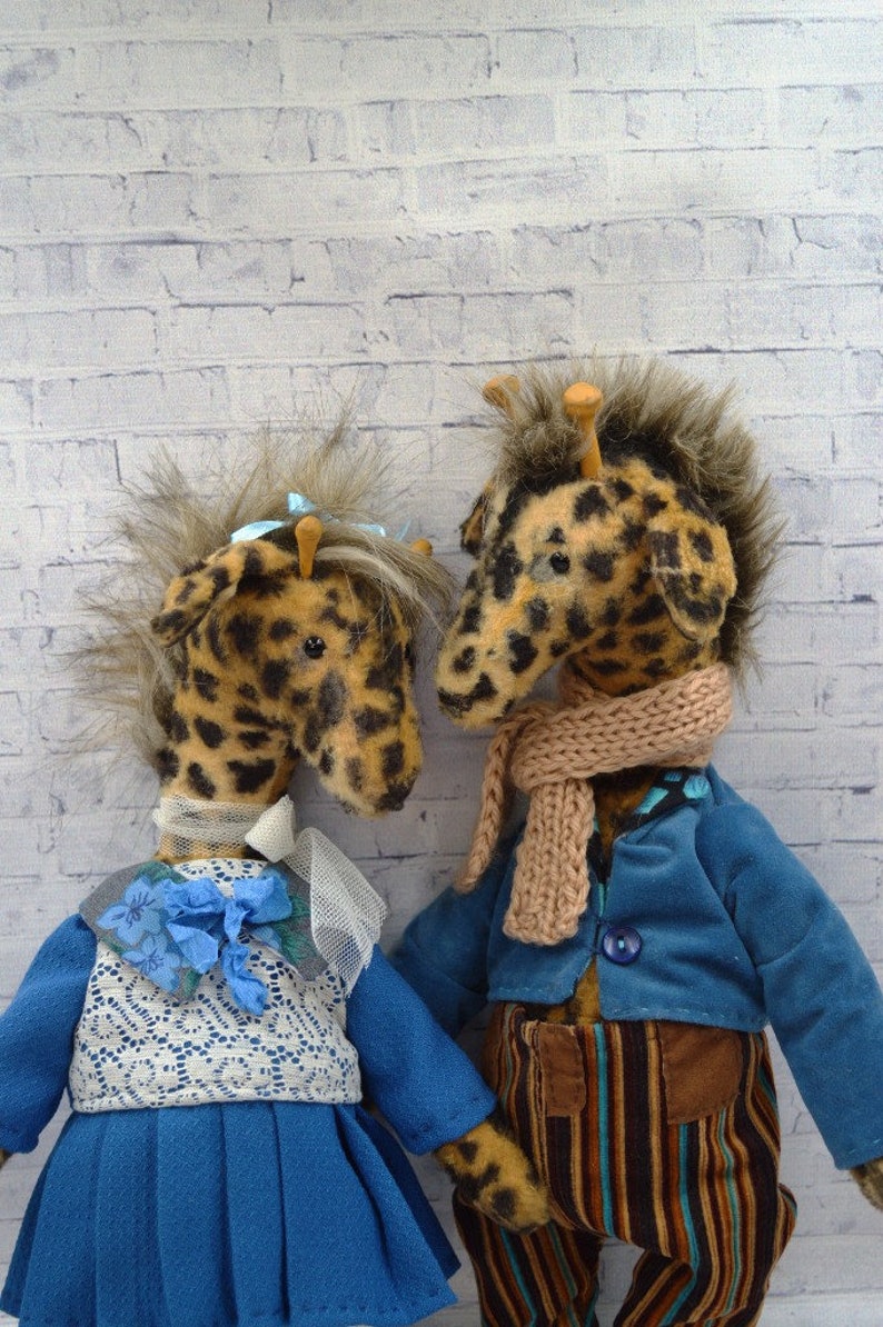 Puede incluir: Dos mu&ntilde;ecos de peluche de jirafa con pelaje moteado, uno frente al otro. Uno lleva un vestido azul con un chaleco de encaje blanco y una flor azul. El otro lleva una chaqueta azul, pantalones a rayas y una bufanda beige.