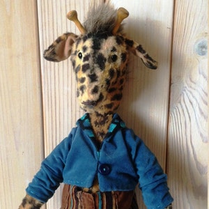Puede incluir: Un peluche de jirafa hecho a mano con cabeza y cuello manchados, que lleva una chaqueta azul y pantalones a rayas. La jirafa tiene peque&ntilde;os cuernos de madera y un mech&oacute;n de pelo gris. Un juguete de peluche &uacute;nico y hecho a mano.