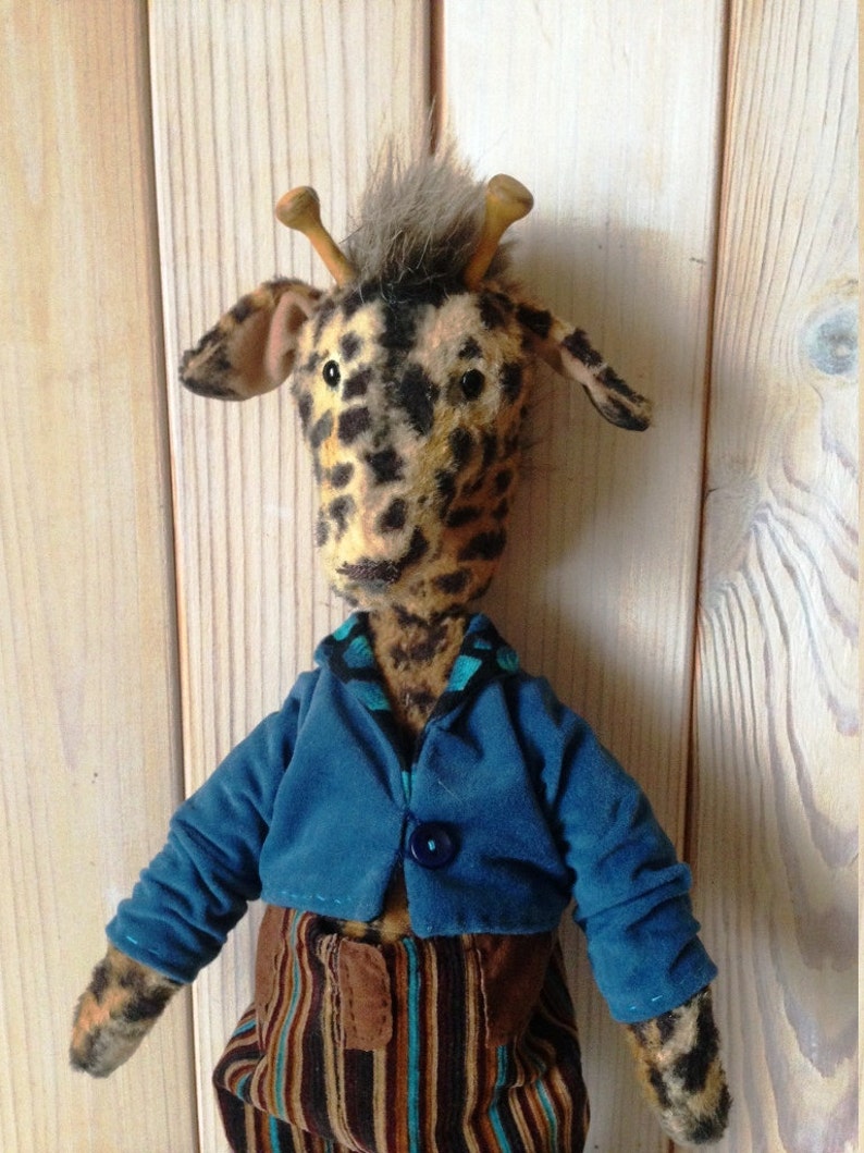 Puede incluir: Un peluche de jirafa hecho a mano con cabeza y cuello manchados, que lleva una chaqueta azul y pantalones a rayas. La jirafa tiene peque&ntilde;os cuernos de madera y un mech&oacute;n de pelo gris. Un juguete de peluche &uacute;nico y hecho a mano.