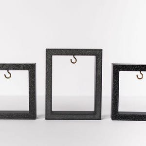 May include: Three square, dark gray frames with a textured finish. Each frame has a small, bronze-colored hook suspended from the top. The frames are arranged in a row, varying in size, against a white background.